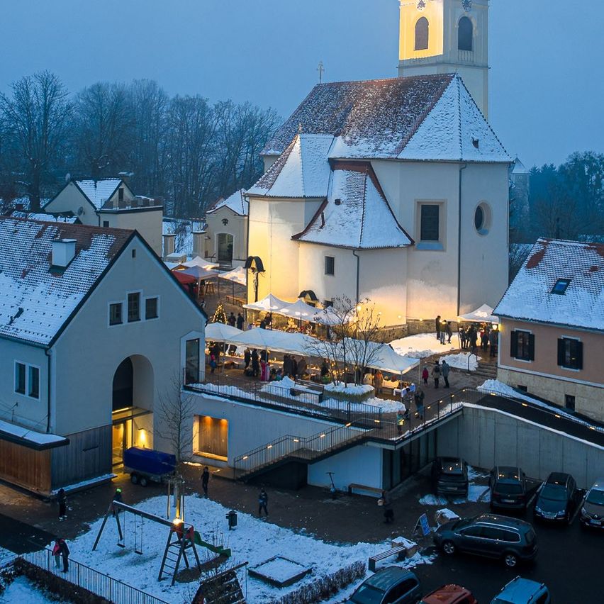 Auf einem verschneiten Hügel steht eine Kirche mit einem Turm, die beleuchtet ist. In der Nähe sind mehrere Gebäude schneebedeckt, und vor ihnen sind Autos geparkt. Der Himmel ist bedeckt.