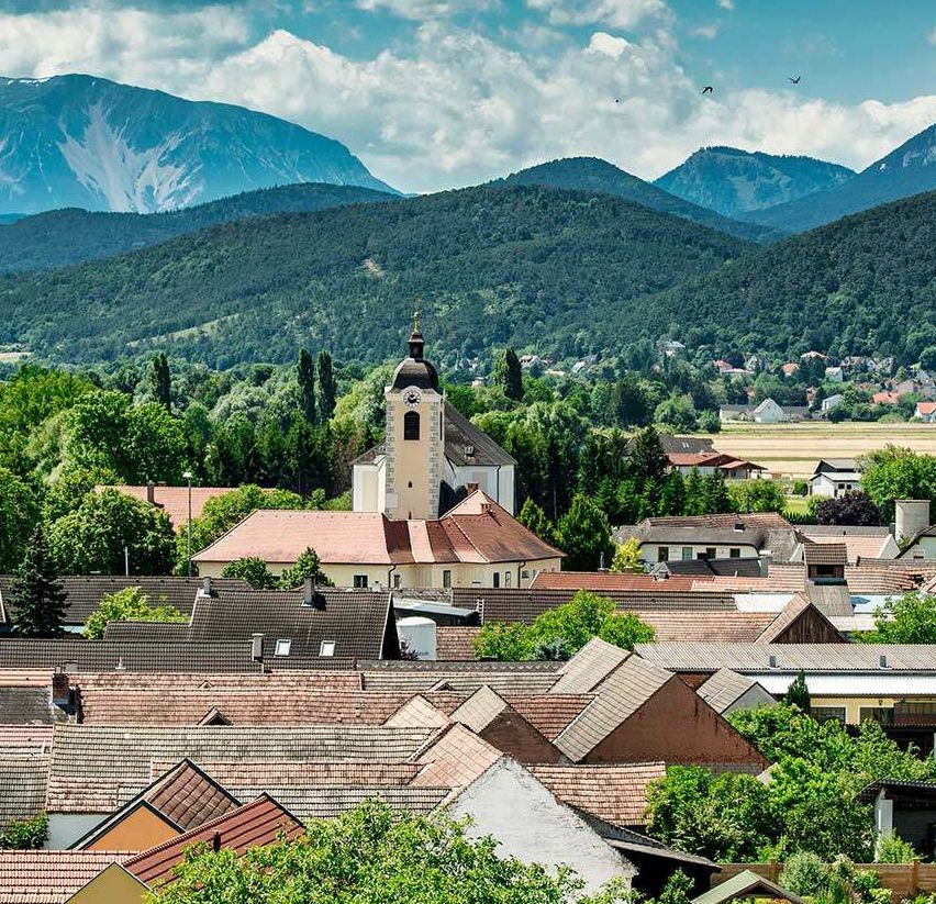 Luftaufnahme einer Stadt umgeben von Bäumen und Bergen. Die Stadt zeigt zahlreiche Gebäude mit braunen Dächern. Eine auffällige Kirche mit Turm steht im Zentrum. Der Himmel ist blau mit verstreuten Wolken.