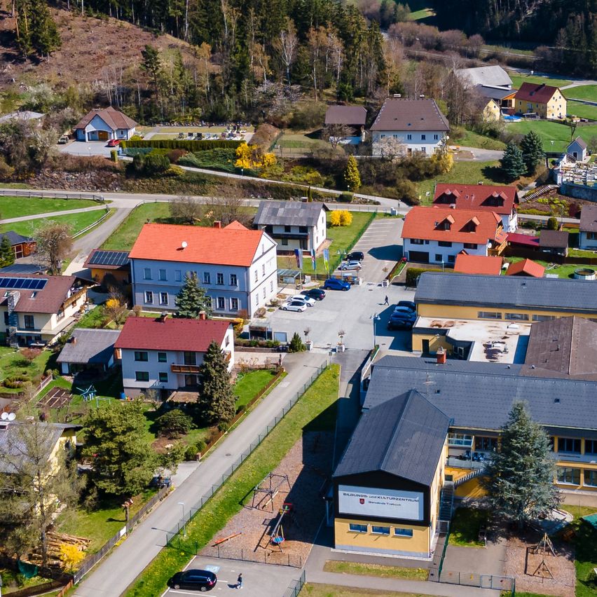 Eine Luftaufnahme eines Dorfes mit mehreren Häusern, einige mit Solarmodulen, umgeben von Grünflächen und Bäumen. Eine Straße mit geparkten Autos und einem Spielplatz ist im Vordergrund zu sehen.