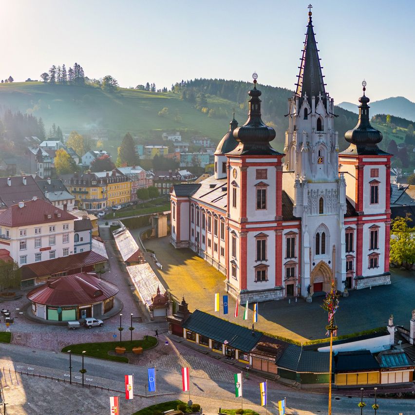 Eine Luftaufnahme einer Stadt mit einer großen Kirche in der Mitte, umgeben von bunten Flaggen und Gebäuden, unter einem klaren blauen Himmel.