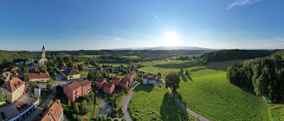 Eine Luftaufnahme eines ländlichen Gebiets mit grünen Feldern und roten Gebäuden unter einem hellen Sonnenschein, mit Bäumen und einem Weg im Vordergrund.