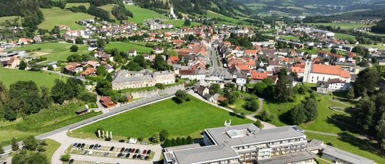 Luftaufnahme eines Dorfes mit einem großen Hotel und einem Schwimmbad. Das Dorf ist von grünen Feldern und Bergen umgeben. Das Hotel verfügt über ein Schwimmbad und einen Parkplatz mit vielen Autos.