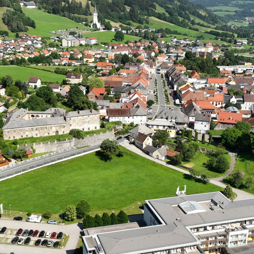 Luftaufnahme eines Dorfes mit einem großen Hotel und einem Schwimmbad. Das Dorf ist von grünen Feldern und Bergen umgeben. Das Hotel verfügt über ein Schwimmbad und einen Parkplatz mit vielen Autos.