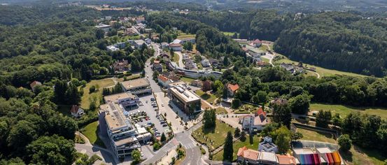 Luftaufnahme einer Stadt mit vielen Gebäuden, geparkten Autos und einer kurvenreichen Straße, umgeben von Bäumen und Gras. Berge in der Ferne.
