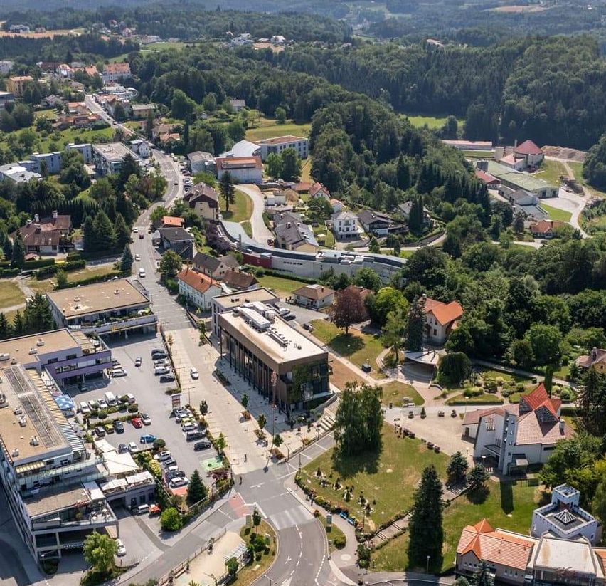 Luftaufnahme einer Stadt mit vielen Gebäuden, geparkten Autos und einer kurvenreichen Straße, umgeben von Bäumen und Gras. Berge in der Ferne.