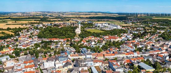 Luftaufnahme einer Stadt mit vielen Gebäuden, einer Kirche und Bäumen vor dem Hintergrund eines blauen Himmels und Windräder in der Ferne.