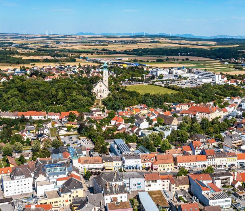 Luftaufnahme einer Stadt mit vielen Gebäuden, einer Kirche und Bäumen vor dem Hintergrund eines blauen Himmels und Windräder in der Ferne.