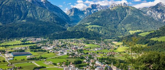 Luftaufnahme eines Dorfes in einem Tal, umgeben von üppigen grünen Feldern und majestätischen Bergen unter einem blauen Himmel mit verstreuten Wolken.