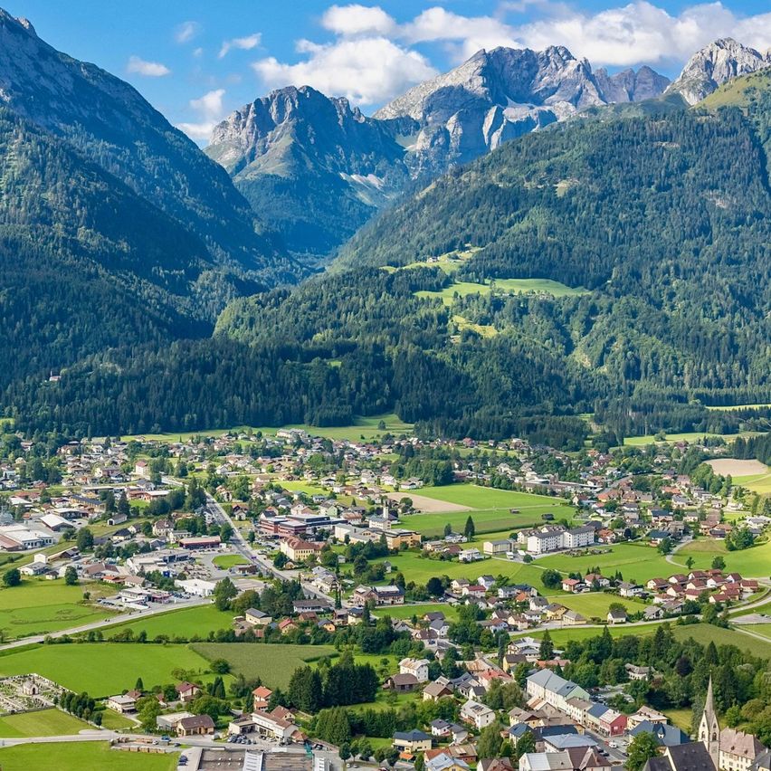 Luftaufnahme eines Dorfes in einem Tal, umgeben von üppigen grünen Feldern und majestätischen Bergen unter einem blauen Himmel mit verstreuten Wolken.
