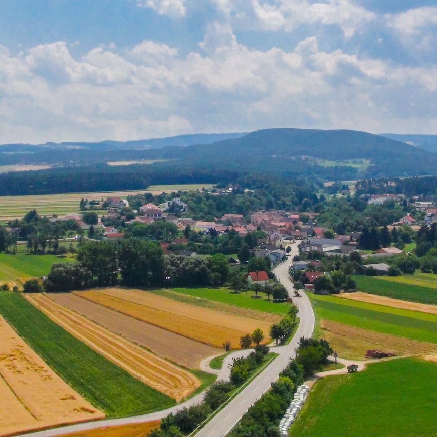 Luftaufnahme einer ländlichen Landschaft mit Feldern, Häusern und einer kurvenreichen Straße unter einem teilweise bewölkten Himmel.