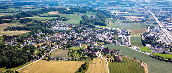 Ein Luftbild einer Stadt, umgeben von üppigen grünen Feldern und Bäumen. Die Stadt besteht aus verschiedenen Gebäuden, einige mit roten Dächern. Die Landschaft umfasst eine Mischung aus Wohngebieten und landwirtschaftlichen Flächen.