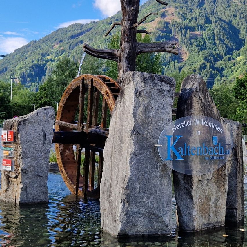Ein Brunnen in der Mitte eines Wasserspiels mit einem Baum darauf und einem Berg im Hintergrund.