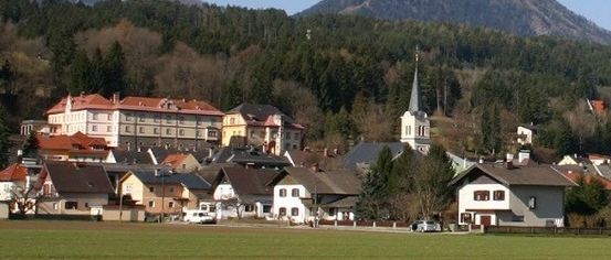 Ein kleines Dorf in den Bergen mit einer Kirche, umgeben von Bäumen und Grünflächen. Im Hintergrund sind schneebedeckte Berge zu sehen.