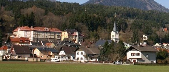 Ein kleines Dorf in den Bergen mit einer Kirche, umgeben von Bäumen und Grünflächen. Im Hintergrund sind schneebedeckte Berge zu sehen.