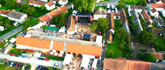 Ein Luftbild eines Dorfes mit Gebäuden, einem grünen Feld und einer Versammlung in der Mitte. Viele Autos sind geparkt und Zelte sind aufgebaut. Bäume umgeben das Gebiet.