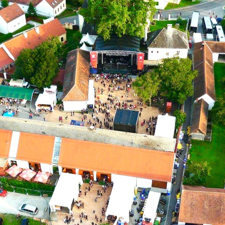 Ein Luftbild eines Dorfes mit Gebäuden, einem grünen Feld und einer Versammlung in der Mitte. Viele Autos sind geparkt und Zelte sind aufgebaut. Bäume umgeben das Gebiet.