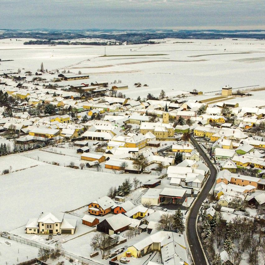 Luftaufnahme eines kleinen Ortes im Winter mit schneebedeckten Häusern, einer kurvenreichen Straße und verschneiten Feldern. Der Ort hat eine Mischung aus gelben, weißen und blauen Häusern.