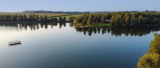 Eine friedliche Landschaft zeigt einen See mit klaren Reflexionen, umgeben von üppigem Grün und einem kleinen Haus. Bäume rahmen die Szene ein, und ein Bootsanleger erstreckt sich ins Wasser.