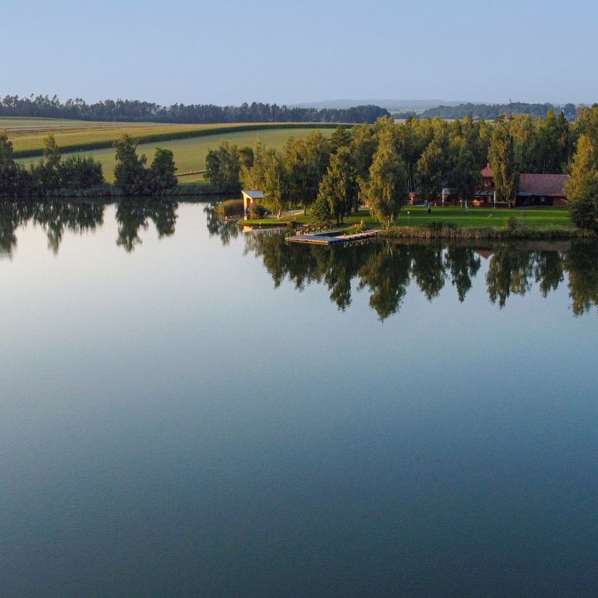 Eine friedliche Landschaft zeigt einen See mit klaren Reflexionen, umgeben von üppigem Grün und einem kleinen Haus. Bäume rahmen die Szene ein, und ein Bootsanleger erstreckt sich ins Wasser.