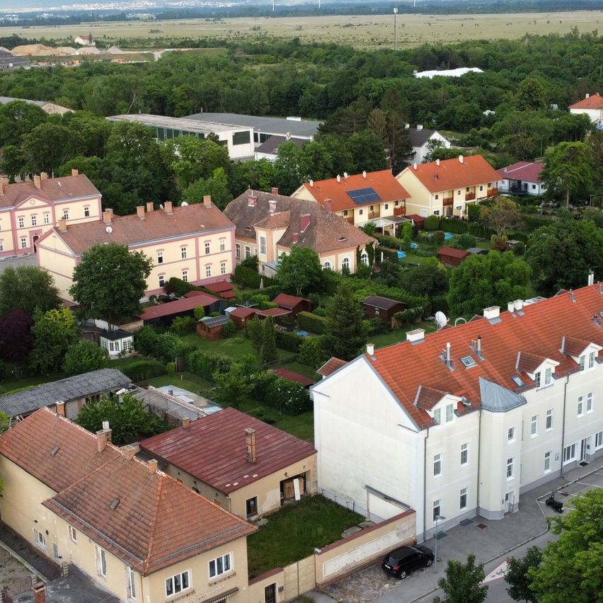 Luftaufnahme eines Vororts mit verschiedenen Gebäuden, einige mit Solarpanelen, und einem auf der Straße geparkten Auto. Das Gebiet ist von Bäumen und Grünflächen umgeben.