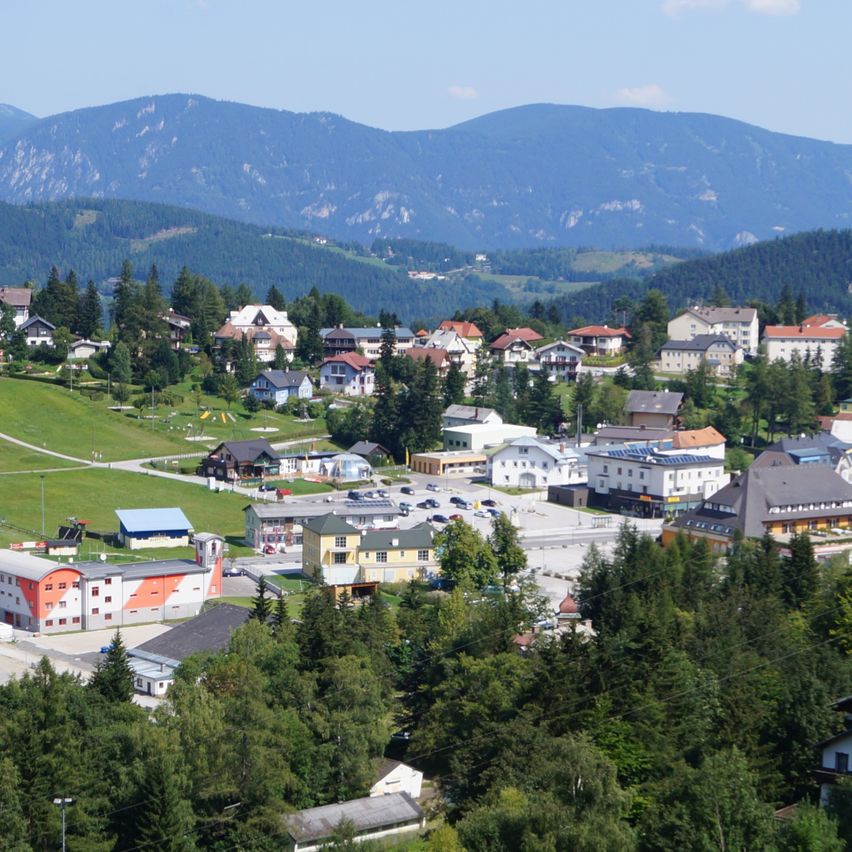 Luftaufnahme einer kleinen Stadt, eingebettet zwischen Bergen und umgeben von üppigem Grün und Bäumen. Die Stadt hat zahlreiche Gebäude und Häuser in verschiedenen Farben. Der Himmel ist klar und blau.