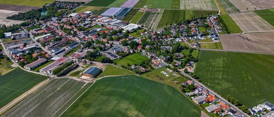 Luftaufnahme einer Stadt mit verschiedenen Gebäuden und grünen Feldern, umgeben von landwirtschaftlichen Flächen und einem klaren blauen Himmel.