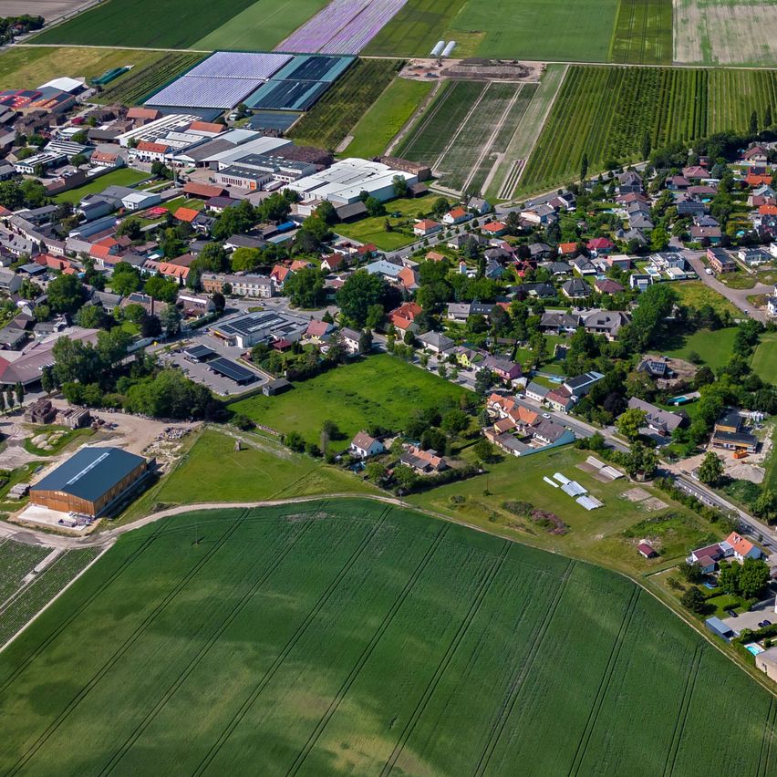 Luftaufnahme einer Stadt mit verschiedenen Gebäuden und grünen Feldern, umgeben von landwirtschaftlichen Flächen und einem klaren blauen Himmel.