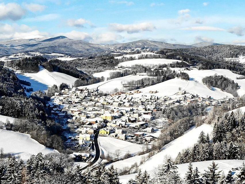 Luftaufnahme eines verschneiten Bergdorfes mit Häusern und Bäumen, die von Schnee bedeckt sind, unter einem klaren blauen Himmel.
