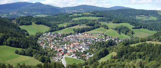Luftaufnahme eines Dorfes in Österreich, umgeben von grünen Hügeln und Bergen, mit Häusern und einer Kirche sichtbar.
