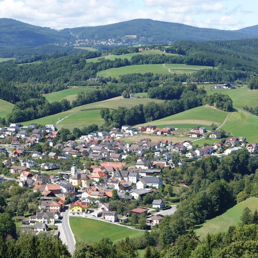 Luftaufnahme eines Dorfes in Österreich, umgeben von grünen Hügeln und Bergen, mit Häusern und einer Kirche sichtbar.