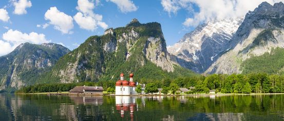 Ein malerischer Alpensee mit einer kleinen Kirche in der Mitte, umgeben von üppigem Grün und majestätischen Bergen unter einem blauen Himmel mit weißen Wolken.