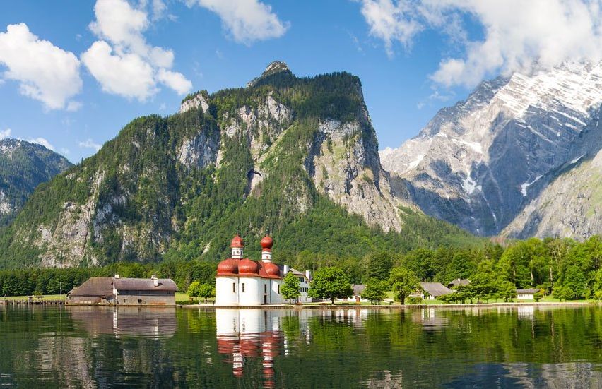 Ein malerischer Alpensee mit einer kleinen Kirche in der Mitte, umgeben von üppigem Grün und majestätischen Bergen unter einem blauen Himmel mit weißen Wolken.