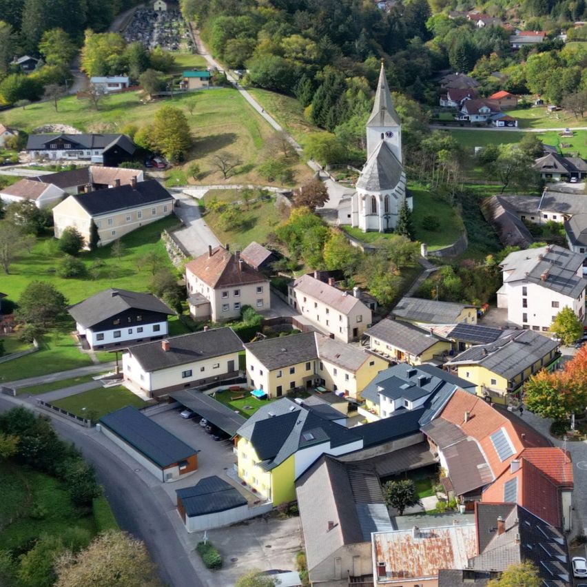 Ein Luftbild eines malerischen Dorfes mit Häusern, einer Kirche und grünen Landschaften unter einem bewölkten Himmel.