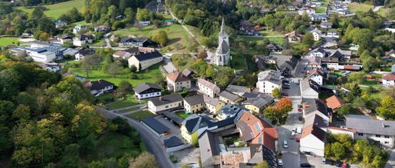 Ein Luftbild eines malerischen Dorfes mit Häusern, einer Kirche und grünen Landschaften unter einem bewölkten Himmel.