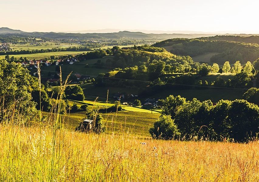 Eine ruhige ländliche Landschaft mit einem Dorf, grünen Hügeln und einem Traktor. Der Himmel ist klar, und die Sonne geht hinter den Hügeln unter.