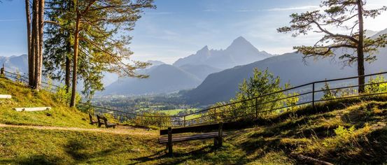 Ein friedlicher Bergblick mit zwei Bänken auf einem Grashügel, der einen Blick auf ein Tal und einen klaren blauen Himmel bietet.