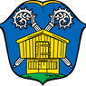Das Wappen zeigt ein gelbes Haus auf blauem Hintergrund mit zwei weißen Zahnrädern darüber. Das Haus hat eine Tür und zwei Fenster.