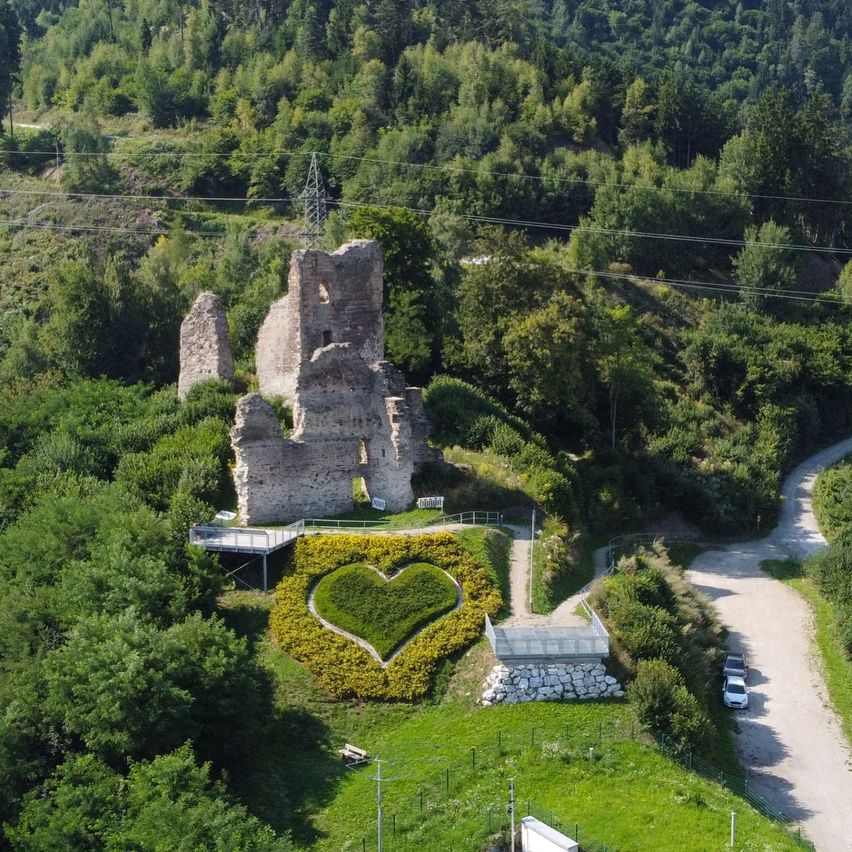 Ein Luftbild von einem Hügel mit einer zerstörten Burg, einem herzförmigen Garten und einem gewundenen Pfad. Bäume umgeben die Burg, und ein Auto ist in der Nähe geparkt.