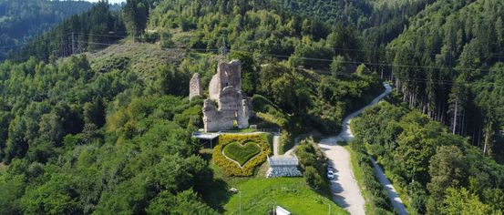 Ein Luftbild von einem Hügel mit einer zerstörten Burg, einem herzförmigen Garten und einem gewundenen Pfad. Bäume umgeben die Burg, und ein Auto ist in der Nähe geparkt.