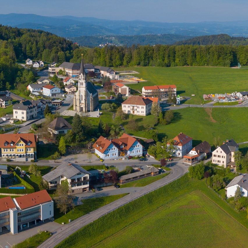 Ein Luftbild eines kleinen Dorfes auf dem Land mit Häusern und einer Kirche. Das Dorf ist von grünen Feldern und Bäumen umgeben. Es gibt eine Straße, die zum Dorf führt.
