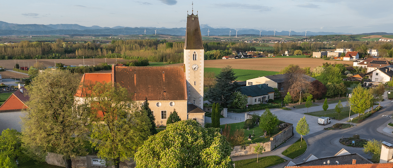 Luftaufnahme einer Kirche mit Turm und Uhrenturm. Umgeben von einem grünen Feld, Häusern und Bäumen. In der Ferne Berge und Windräder.