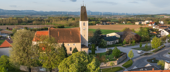 Luftaufnahme einer Kirche mit Turm und Uhrenturm. Umgeben von einem grünen Feld, Häusern und Bäumen. In der Ferne Berge und Windräder.