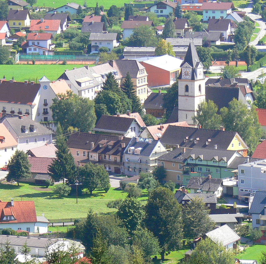Luftaufnahme einer kleinen Stadt mit mehreren Häusern, einem Glockenturm und üppiger Grünfläche. Bäume und Felder umgeben die Stadt.