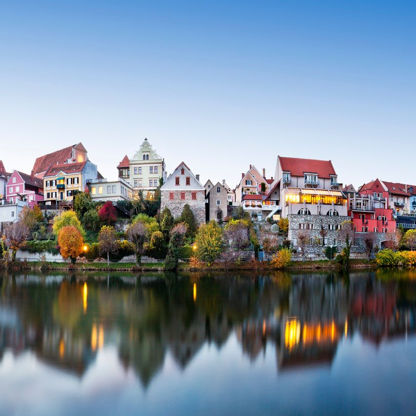 Ein malerischer Blick auf eine historische Stadt mit farbenfrohen Gebäuden, die sich im Wasser spiegeln, unter einem klaren blauen Himmel.