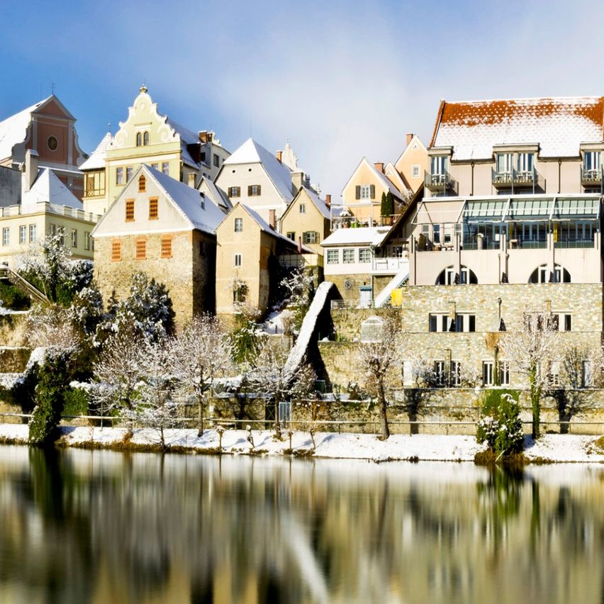 Eine ruhige Stadt mit schneebedeckten Dächern spiegelt sich im ruhigen Wasser unter einem klaren Himmel.
