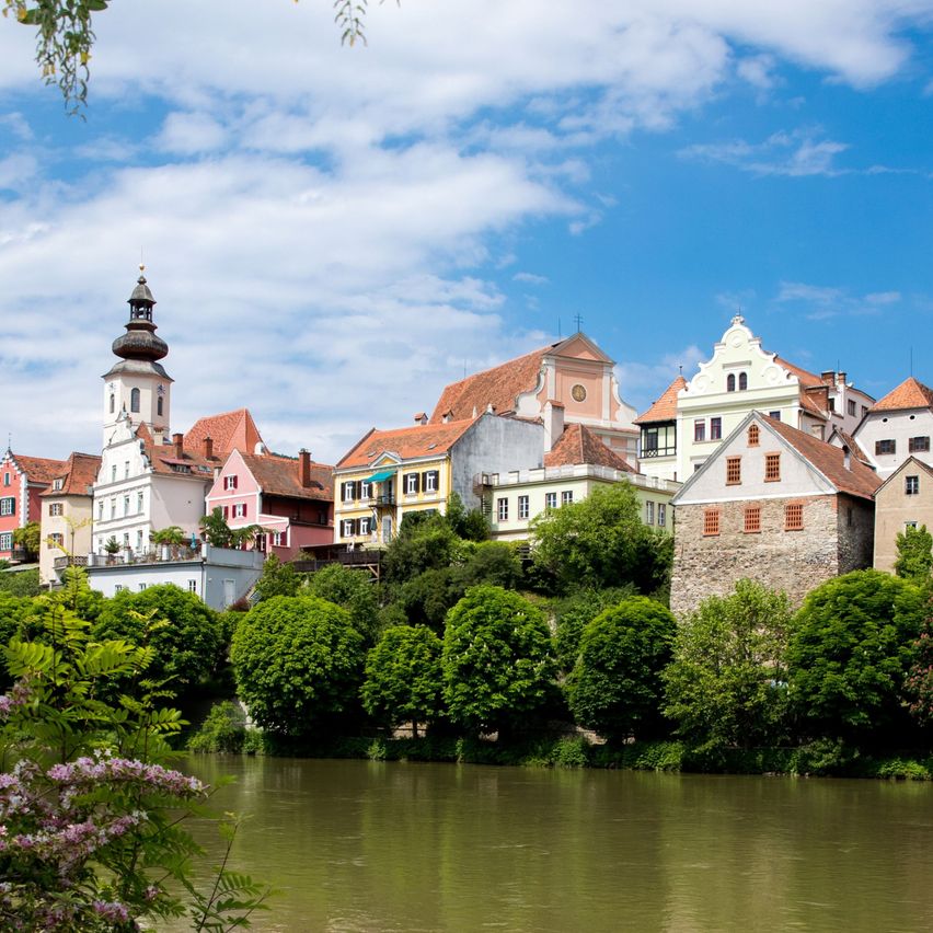 Ein Dorf mit farbenfrohen Gebäuden, eingebettet zwischen üppigem Grün und einem Fluss unter einem blauen Himmel mit Wolken.