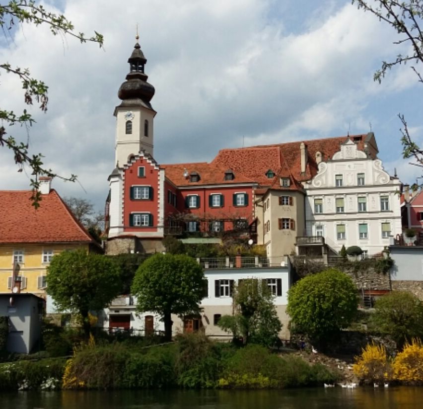 Ein malerischer Blick auf Gebäude mit roten Dächern und einem Glockenturm vor einem blauen Himmel mit Wolken, gelegen an einem Fluss mit Enten und grünen Bäumen.