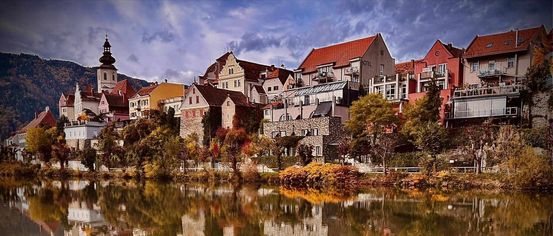 Ein malerisches Dorf mit farbenfrohen Gebäuden, die sich im ruhigen Fluss unter einem bewölkten Himmel spiegeln.