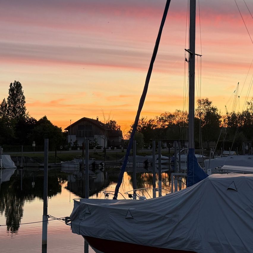 Bei Sonnenuntergang ist ein mit einer Plane bedecktes Boot in einer Marina verankert, mit einem farbenfrohen Himmel. Bäume und Häuser sind in der Ferne sichtbar.