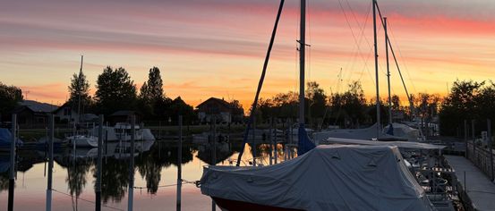 Bei Sonnenuntergang ist ein mit einer Plane bedecktes Boot in einer Marina verankert, mit einem farbenfrohen Himmel. Bäume und Häuser sind in der Ferne sichtbar.